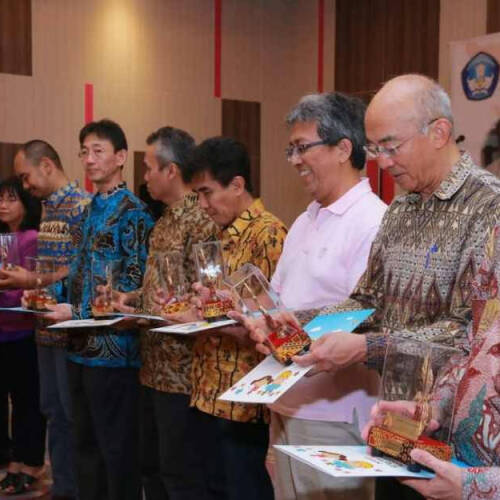 A group of people stands in a row at an indoor event, each holding a clear trophy and a colorful certificate or booklet. They are dressed in traditional and semi-formal batik shirts, with warm expressions on their faces, suggesting a recognition or award.